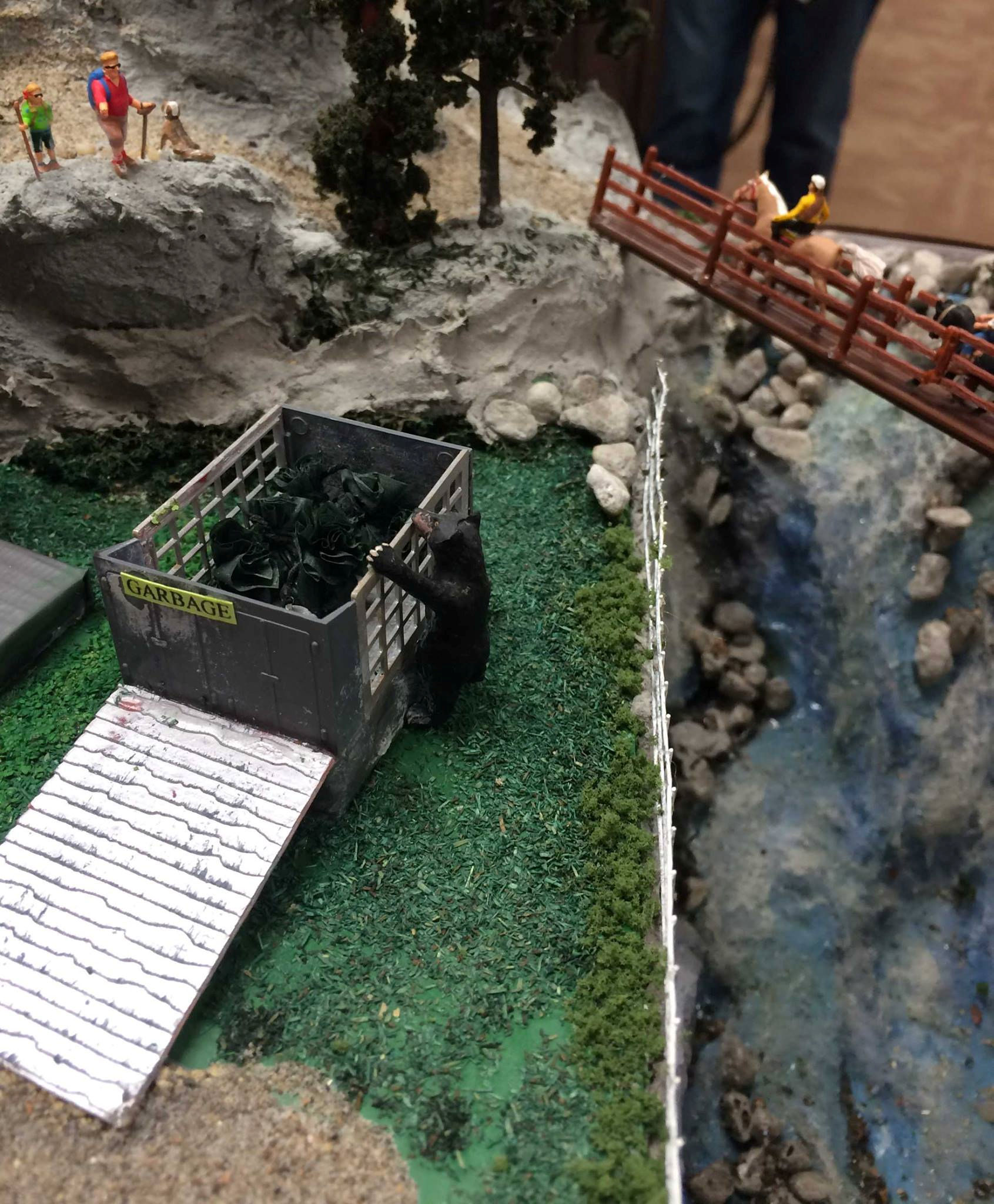Model train layout with a black bear in a garbage bin, hikers, and horseback riders at Oshawa Pine Ridge Model Railroaders Club.