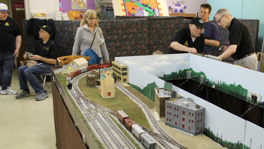 Visitors and club members engage with a model railroad display featuring buildings, tracks, and miniature trains at a club event.