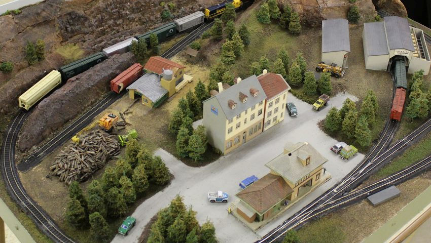 Miniature model train layout with a town, railway tracks, a lumber yard, and industrial buildings.