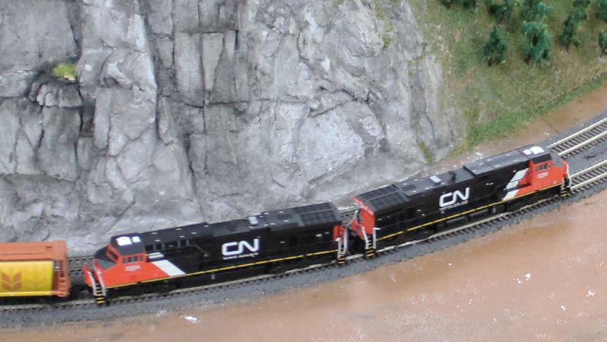 Model train with two Canadian National locomotives pulling freight cars along a curved track near a rocky landscape.