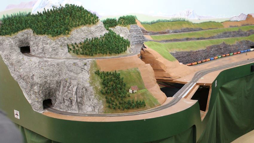 Model railroad with mountain tunnels, curved tracks, and a freight train moving through a detailed landscape.