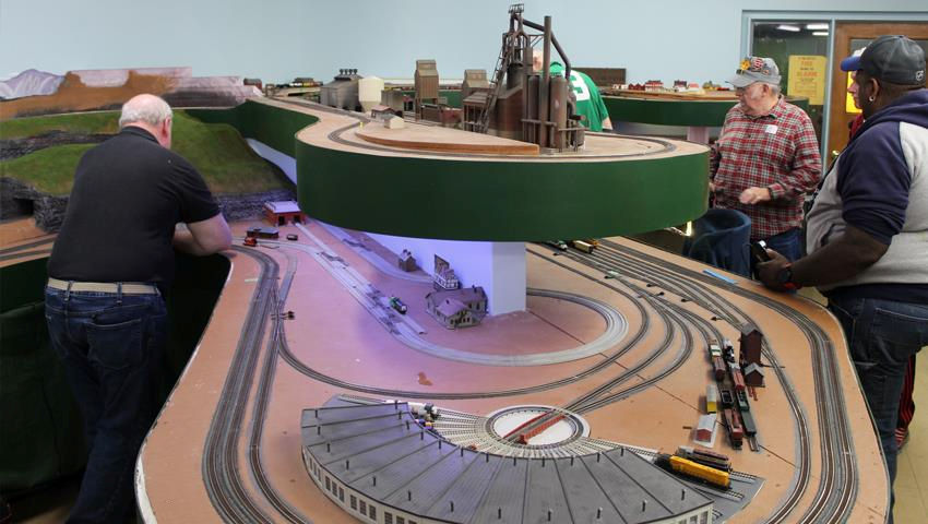 Model train layout with a turntable, multi-track system, and an elevated industrial area, surrounded by railroad enthusiasts.