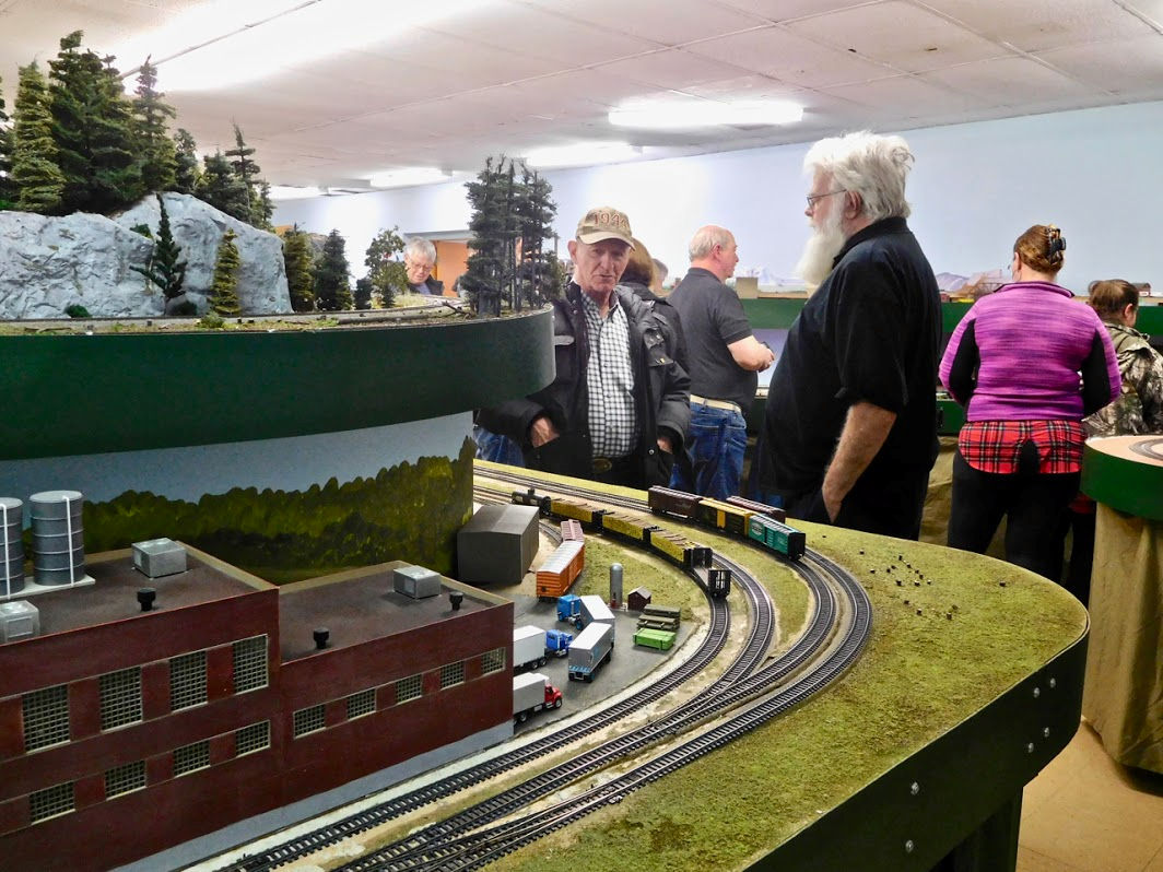 Model railroad enthusiasts observing an industrial-themed layout with train tracks, freight cars, and a factory setting.