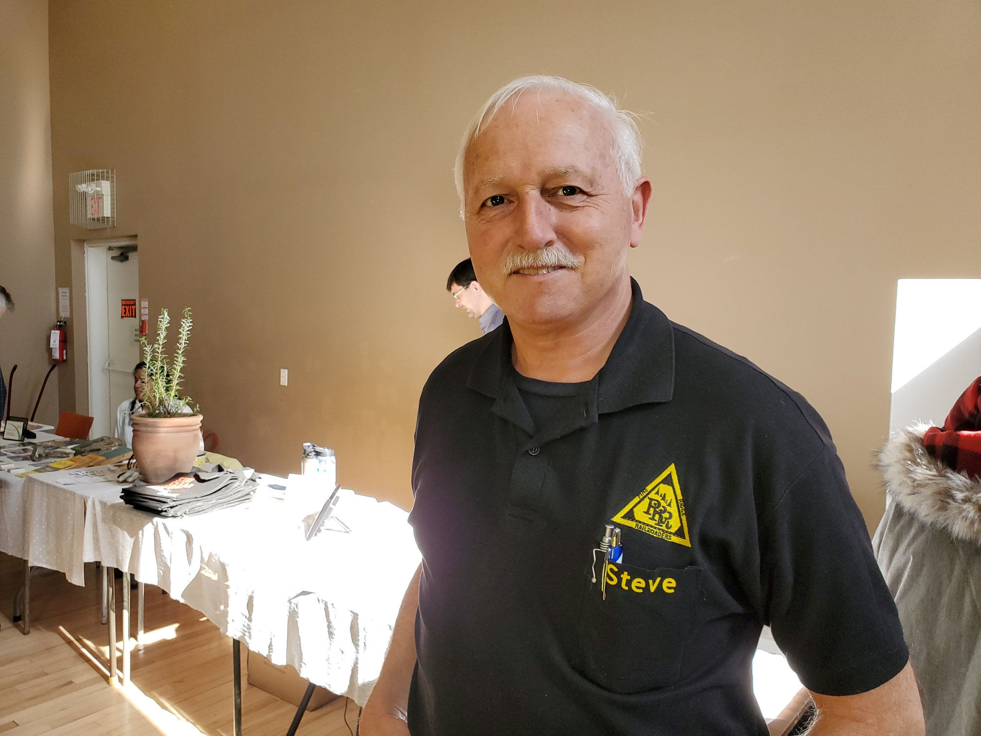 A smiling man wearing a Pine Ridge Model Railroaders Club shirt, standing at an event with tables and displays in the background.