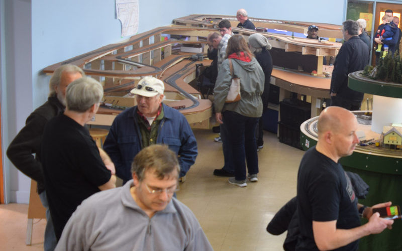 A group of model railroaders and visitors engaging with an expansive model train layout featuring scenic landscapes and train tracks.