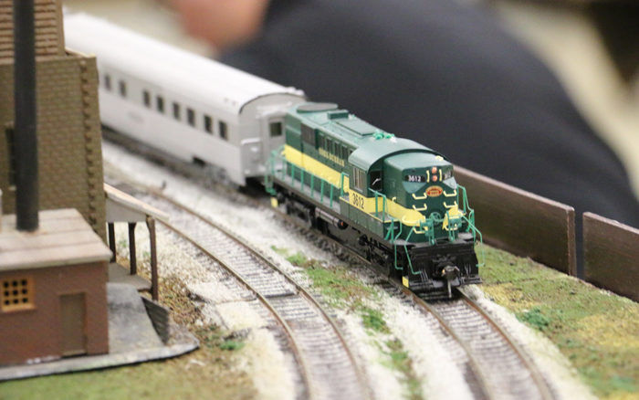 Green and yellow model locomotive pulling a passenger car on a model railroad layout.