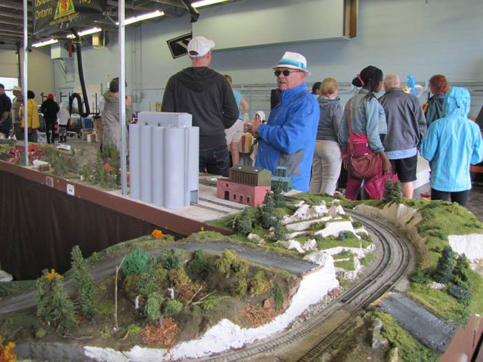 Crowd gathered around an intricate model railroad display featuring a curved railway track, industrial buildings, and scenic landscapes at a train