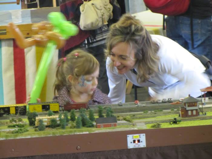 A smiling mother and her young daughter admire a model train layout featuring miniature trees, tracks, and buildings at a train exhibition