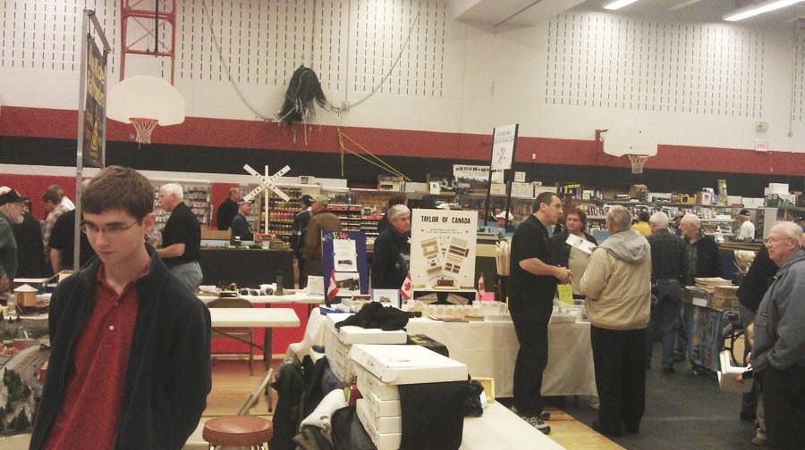 Attendees browsing model train vendors and displays at an indoor model railway exhibition, featuring various booths and hobbyist merchandise.