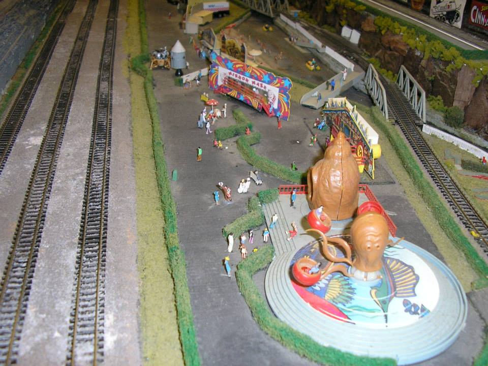 Miniature amusement park with fairground rides, detailed figures, and train tracks in a model railroad display.