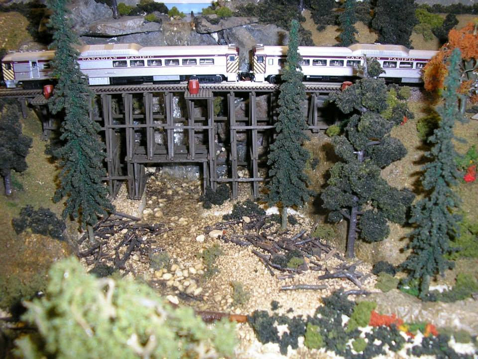 Model train crossing a wooden trestle bridge surrounded by trees and rocky terrain in a detailed railroad layout.