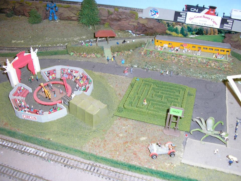 Model railroad layout with a circus performance, a hedge maze, a futuristic robot, and a train passing on an elevated track.