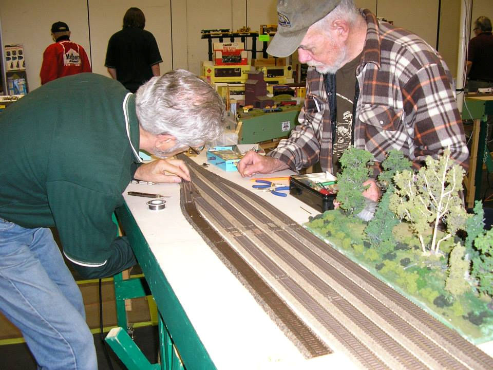 Two model railroaders installing track on a model train layout, using tools for precision detailing.