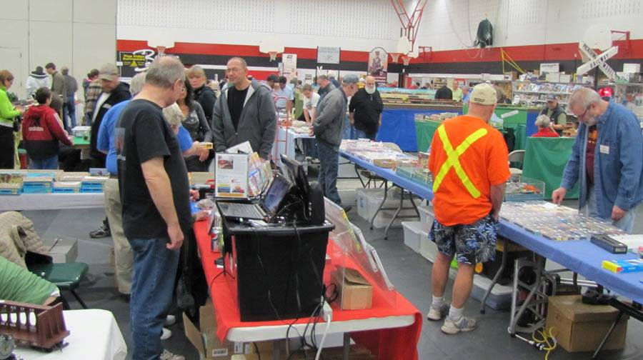 Train show attendees browsing model railroad displays and vendor tables in a large indoor venue.
