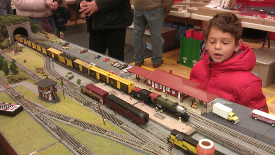 Young boy in a red coat observing a model train station with freight cars, locomotives, and a tunnel.