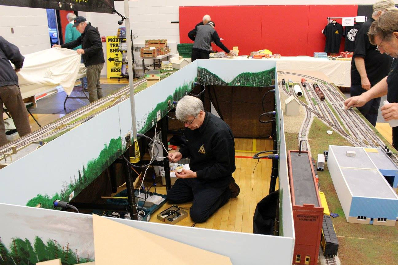 Man kneeling under a model train layout working on electrical components, with other hobbyists setting up tables in the background.