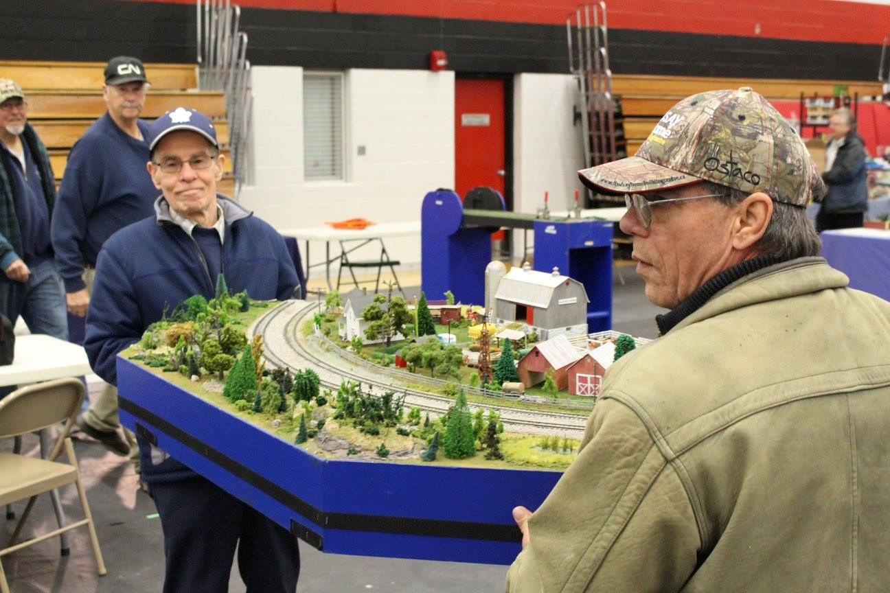 Two men carrying a modular model train layout with a curved railway, trees, and farm buildings at a model railroad exhibition.