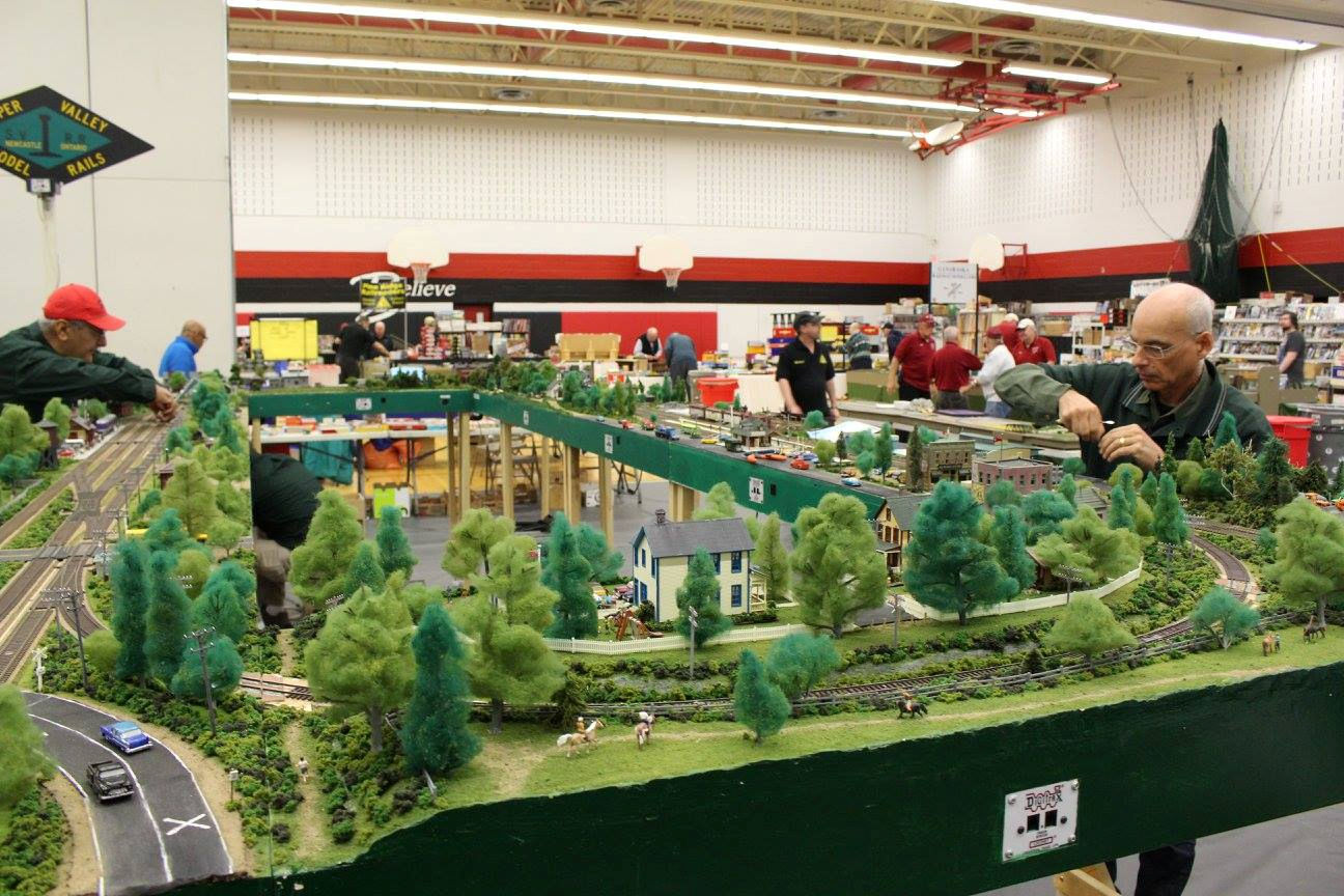 Model railroad enthusiasts working on a large, detailed train layout with trees, buildings, roads, and multiple tracks at an exhibition.
