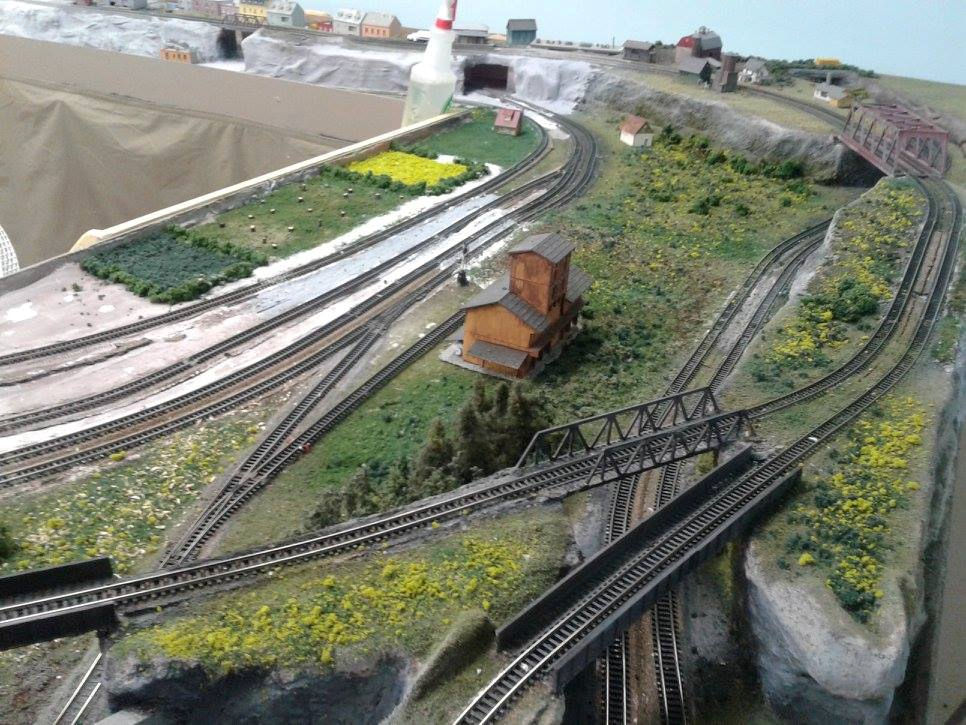 Model train layout with multiple tracks, a wooden building, a steel bridge, and realistic terrain with grass and trees.