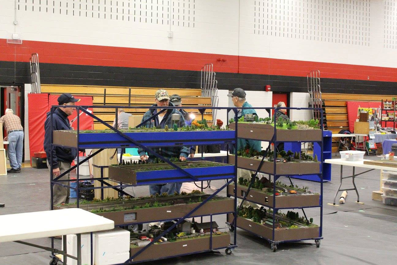 A group of hobbyists handling modular model railroad sections stored on wheeled racks at an indoor event.