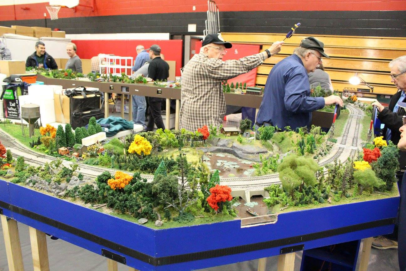 Model railroaders adjusting a train layout with colorful trees, a bridge, and a detailed landscape at an exhibition.