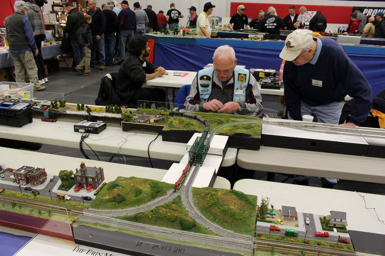 Two men working on a modular model train setup with a bridge and miniature town.