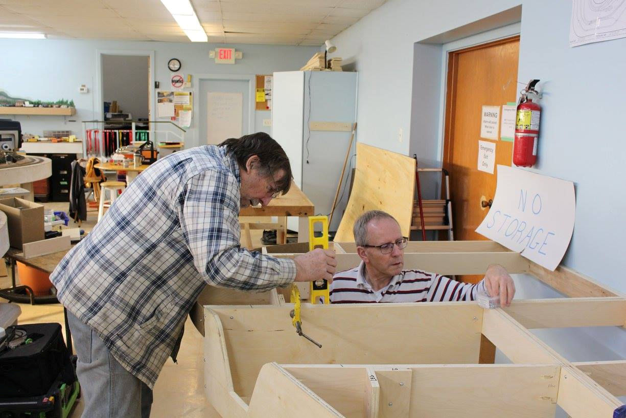 Two model railroad enthusiasts assembling a wooden train layout framework at the Pine Ridge Model Railroaders Club.