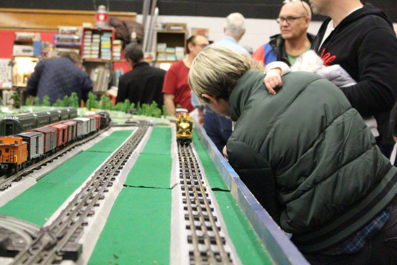 Close-up of a model train approaching on the track with a visitor and child watching attentively at a model railroad event.