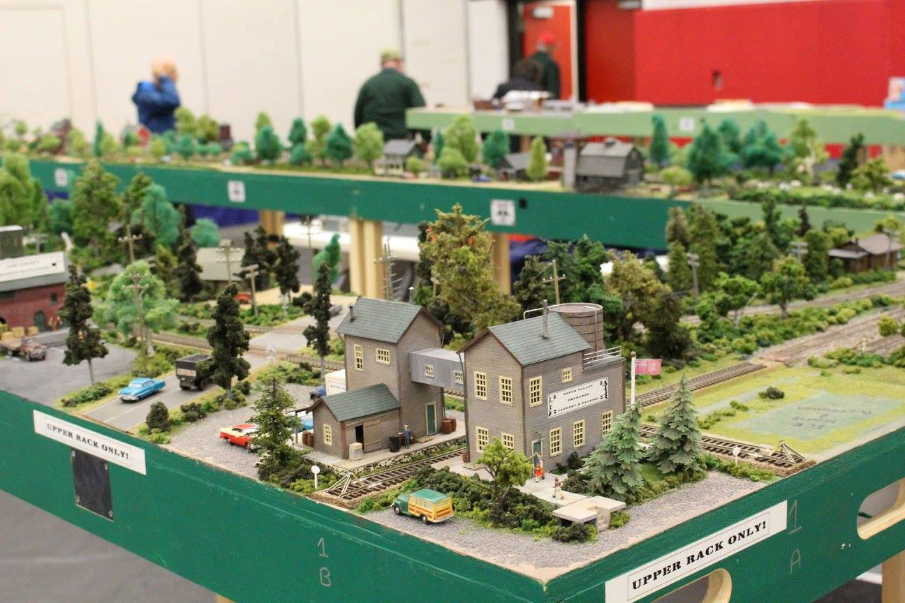 Model railroad layout with an industrial building, trees, vehicles, and a railway track, part of a larger modular train display.
