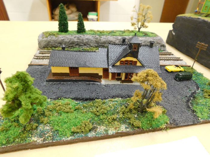 A yellow and black model train station with surrounding greenery, trees, and a roadway.