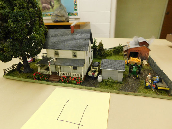 A miniature model of a two-story farmhouse with a barn, a tractor, and small figures in a farm setting